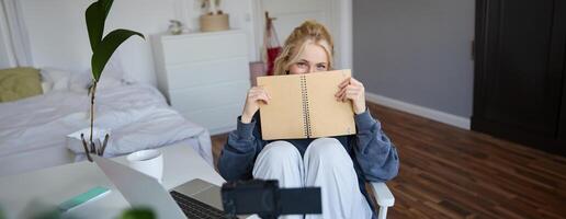 retrato do fofa loiro mulher, senta dentro frente do digital Câmera e computador portátil dentro dela sala, cobre face com caderno Diário, registros do ela mesma para estilo de vida vlog foto