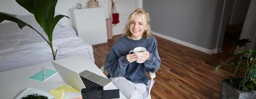 imagem do jovem mulher, social meios de comunicação influenciador, edição dela em computador portátil, senta dentro uma quarto com computador e digital Câmera, bebendo café, sorridente às Câmera foto