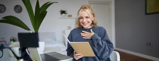 retrato do sorridente jovem fofa mulher, segurando caderno, menina com computador portátil e planejador dentro mãos, senta dentro sala, parece às digital Câmera, cria estilo de vida vlog, fala para público, lê roteiro foto