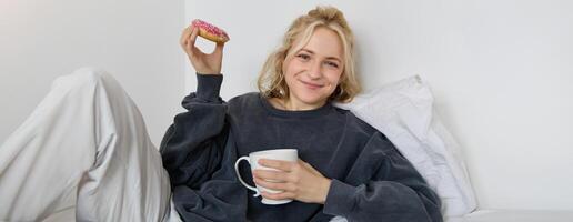 estilo de vida e pessoas conceito. feliz loiro mulher deitado dentro cama com comida, bebendo chá e comendo rosquinha, em repouso às lar, gastos Tempo sozinho, assistindo televisão mostrar foto