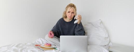 retrato do mulher assistindo triste filme em computador portátil, chorando e limpando lágrimas desligado, comendo rosquinha e bebendo chá foto
