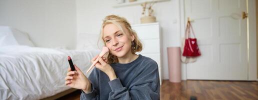 mulher beleza blogueiro, registros do ela mesma sentado dentro uma quarto e Avaliação Maquiagem produtos, coloca em faço acima, detém batom e Cosmético escova dentro mão, usando profissional Câmera para conteúdo criação foto