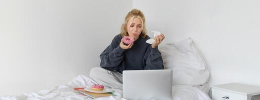 retrato do mulher assistindo triste mostrar em computador portátil, comendo rosquinha e limpando lágrimas com guardanapo, sentado em cama, gastos Tempo às casa enquanto ser chateado foto