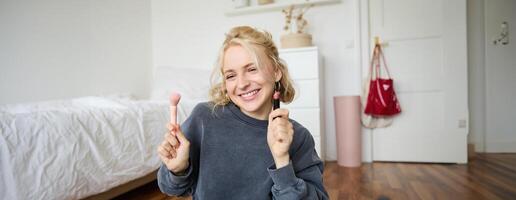 mulher beleza blogueiro, registros do ela mesma sentado dentro uma quarto e Avaliação Maquiagem produtos, coloca em faço acima, detém batom e Cosmético escova dentro mão, usando profissional Câmera para conteúdo criação foto