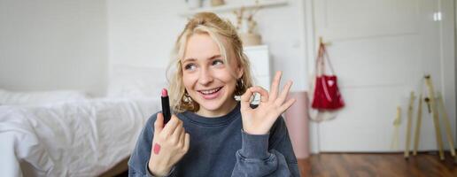 jovem fêmea blogueiro, conteúdo O Criador mostrando batom e OK mão sinal, recomendando beleza produtos para dela público em social meios de comunicação, gravação vlog dentro quarto foto