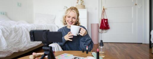 alegre mulher, beleza blogueiro, registros estilo de vida vlog em digital Câmera, fala casualmente, conta uma história para social meios de comunicação seguidores, detém xícara, bebidas chá e senta em chão dentro dela quarto foto