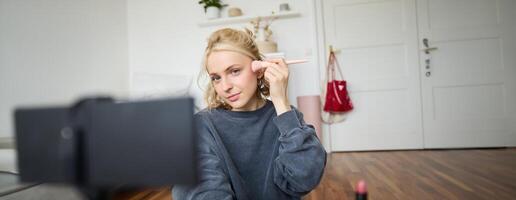 retrato do jovem lindo mulher aplica-se Maquiagem cora com uma escova e registros ela mesma em digital Câmera, cria conteúdo para beleza blog, social meios de comunicação conta foto