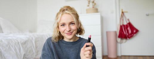 retrato do sorridente lindo mulher dentro dela sala, sentado e mostrando batom, recomendando favorito beleza produtos, conteúdo criador gravação uma do ela mesma para social meios de comunicação blog foto