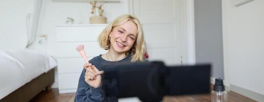 fechar acima retrato do feliz jovem beleza blogueiro, registros estilo de vida vlog dentro dela sala, usando Câmera com estabilizador, mostra Maquiagem escova e cosméticos foto