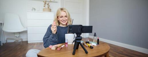 retrato do lindo, sorridente loiro mulher, menina gravação do dela Maquiagem tutorial para social meios de comunicação, vlogger sentado em chão dentro dela sala, usando estabilizador para crio contente, revendo rímel foto