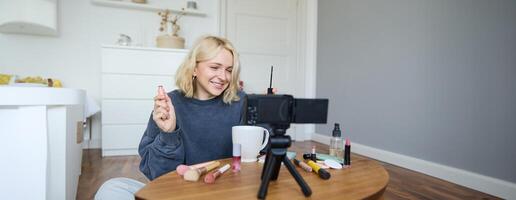 retrato do loiro sorridente mulher registros uma estilo de vida blog, vlogger ou Maquiagem artista gravação para social meios de comunicação, segurando rímel, revendo beleza produtos para seguidores conectados foto