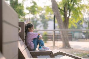 uma pequeno menina senta em uma Banco dentro uma parque foto