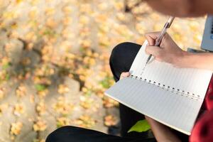 aluna sentado ao ar livre dentro natureza parque escrevendo notas dentro caderno. homem mão com caneta escrevendo às a manhã foto