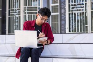 ásia Faculdade aluna estudando em computador portátil às campus ao ar livre parque. homem escrevendo em uma Nota livro e trabalhando em computador portátil. educacional conceito foto