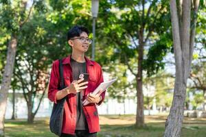 jovem ásia Faculdade aluna usando Smartphone com feliz expressão. uma masculino sorridente enquanto segurando dele telefone e livros às a público parque. cópia de espaço foto