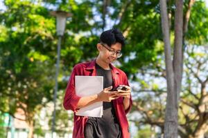 jovem ásia Faculdade aluna usando Smartphone com feliz expressão. uma masculino sorridente enquanto segurando dele telefone e livros às a público parque. cópia de espaço foto