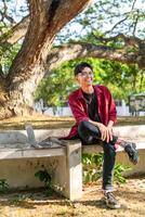 jovem ásia homem sentado ao ar livre com feliz expressão. uma masculino Faculdade aluna sorridente às a público parque foto