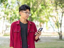 retrato do ásia homem sorridente enquanto segurando dele telefone. homem com vermelho camisa sorridente alegremente às Câmera enquanto usando Smartphone foto