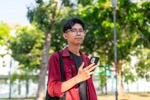 jovem ásia homem usando Smartphone com feliz expressão. uma masculino sorridente enquanto segurando dele telefone às a público parque foto