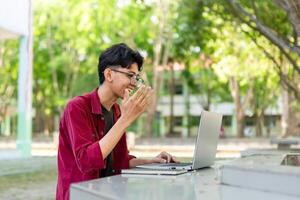 jovem ásia homem sorridente enquanto usando computador portátil para trabalhando e conferência encontro às a manhã. ásia homem tendo uma ligar em computador portátil. conceito do controlo remoto e freelance foto