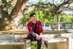 jovem ásia homem sentado ao ar livre com feliz expressão. uma masculino Faculdade aluna sorridente às a público parque foto