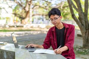 ásia Faculdade aluna estudando em computador portátil às campus ao ar livre parque. homem escrevendo em uma Nota livro e trabalhando em computador portátil. educacional conceito foto