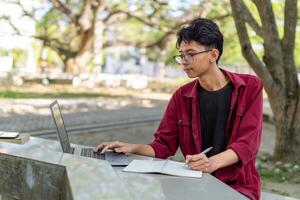 ásia Faculdade aluna estudando em computador portátil às campus ao ar livre parque. homem escrevendo em uma Nota livro e trabalhando em computador portátil. educacional conceito foto