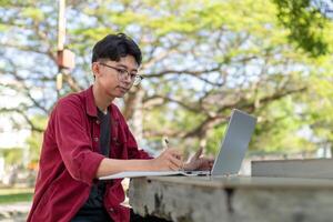 ásia Faculdade aluna estudando em computador portátil às campus ao ar livre parque. homem escrevendo em uma Nota livro e trabalhando em computador portátil. educacional conceito foto