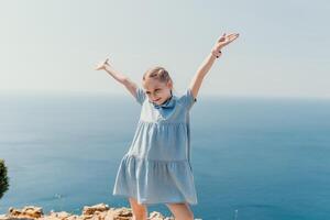 uma jovem menina é em pé em uma praia, vestindo uma azul vestir e segurando dela braços acima dentro a ar. conceito do alegria e liberdade, Como a menina parece para estar desfrutando dela Tempo de a oceano. foto