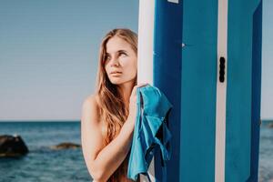 mulher mar e aí. fechar acima retrato do feliz jovem caucasiano mulher com grandes cabelo olhando às Câmera e sorridente. fofa mulher retrato dentro uma azul bikini posando em sup borda dentro a mar foto