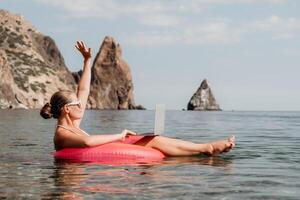 mulher trabalhador autonomo trabalho em computador portátil natação dentro mar em Rosa inflável anel. feliz turista dentro oculos de sol flutuando em inflável rosquinha e trabalhando em computador portátil computador dentro calma oceano. controlo remoto trabalhando qualquer lugar foto