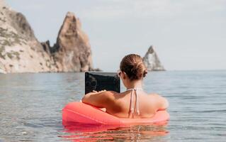 mulher trabalhador autonomo trabalho em computador portátil natação dentro mar em Rosa inflável anel. feliz turista dentro oculos de sol flutuando em inflável rosquinha e trabalhando em computador portátil computador dentro calma oceano. controlo remoto trabalhando qualquer lugar foto