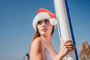 mulher mar e aí. fechar acima retrato do feliz jovem caucasiano mulher com grandes cabelo dentro santa chapéu olhando às Câmera e sorridente. fofa mulher retrato dentro uma branco bikini posando em sup borda dentro a mar foto