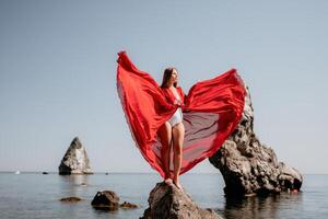 mulher viagem mar. jovem feliz mulher dentro uma grandes vermelho vestir posando em uma de praia perto a mar em fundo do vulcânico rochas, gostar dentro Islândia, partilha viagem aventura viagem foto