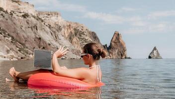 mulher trabalhador autonomo trabalho em computador portátil natação dentro mar em Rosa inflável anel. feliz turista dentro oculos de sol flutuando em inflável rosquinha e trabalhando em computador portátil computador dentro calma oceano. controlo remoto trabalhando qualquer lugar foto