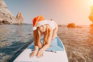 mulher mar e aí. fechar acima retrato do feliz jovem caucasiano mulher com grandes cabelo dentro santa chapéu olhando às Câmera e sorridente. fofa mulher retrato dentro uma branco bikini posando em sup borda dentro a mar foto