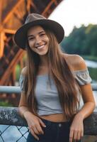 caucasiano mulher por aí 25 anos velho com grandes Castanho cabelo vestindo uma chapéu, uma cinzento Fora do ombro principal, e acessórios, sorridente, com uma ponte e árvores dentro foto