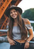 caucasiano mulher por aí 25 anos velho com grandes Castanho cabelo vestindo uma chapéu, uma cinzento Fora do ombro principal, e acessórios, sorridente, com uma ponte e árvores dentro foto