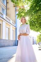 elegante meio era mulher dentro uma branco vintage vestir contra a fundo do histórico edifícios dentro a manhã luz foto