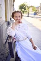 elegante meio era mulher dentro uma branco vintage vestir sentado em uma Banco dentro a manhã cidade foto