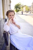 elegante meio era mulher dentro uma branco vintage vestir sentado em uma Banco dentro a manhã cidade foto