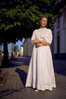 elegante meio era mulher dentro uma branco vintage vestir contra a fundo do histórico edifícios dentro a manhã luz foto