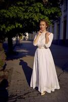 elegante meio era mulher dentro uma branco vintage vestir contra a fundo do histórico edifícios dentro a manhã luz foto