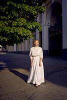 elegante meio era mulher dentro uma branco vintage vestir contra a fundo do histórico edifícios dentro a manhã luz foto