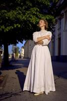 elegante meio era mulher dentro uma branco vintage vestir contra a fundo do histórico edifícios dentro a manhã luz foto