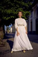 elegante meio era mulher dentro uma branco vintage vestir contra a fundo do histórico edifícios dentro a manhã luz foto