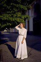 elegante meio era mulher dentro uma branco vintage vestir contra a fundo do histórico edifícios dentro a manhã luz foto