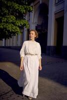 elegante meio era mulher dentro uma branco vintage vestir contra a fundo do histórico edifícios dentro a manhã luz foto