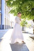 elegante meio era mulher dentro uma branco vintage vestir contra a fundo do histórico edifícios dentro a manhã luz foto