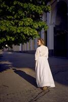 elegante meio era mulher dentro uma branco vintage vestir contra a fundo do histórico edifícios dentro a manhã luz foto